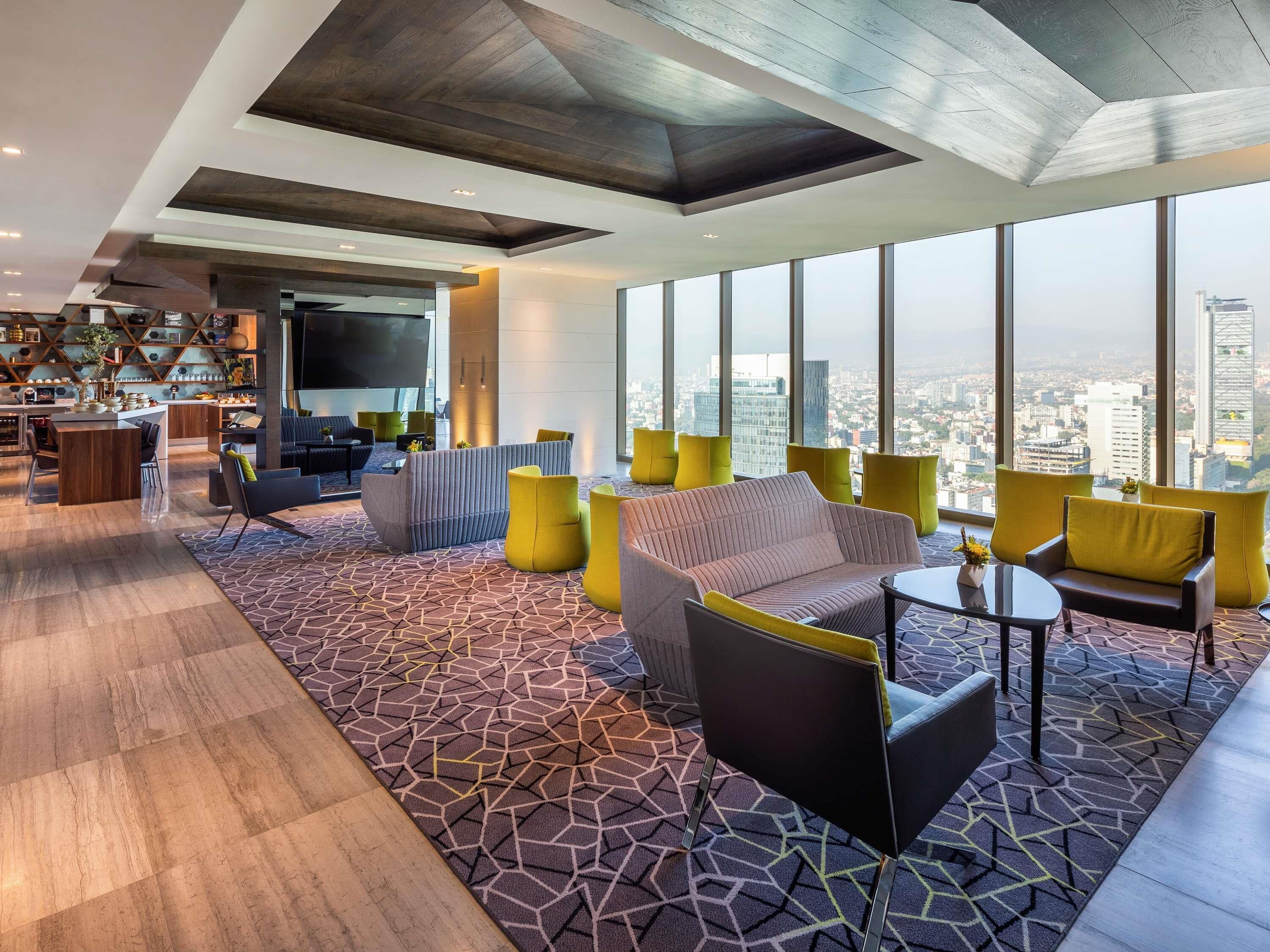 A modern hotel lobby with a large seating area, a bar, and a kitchen visible in the background.