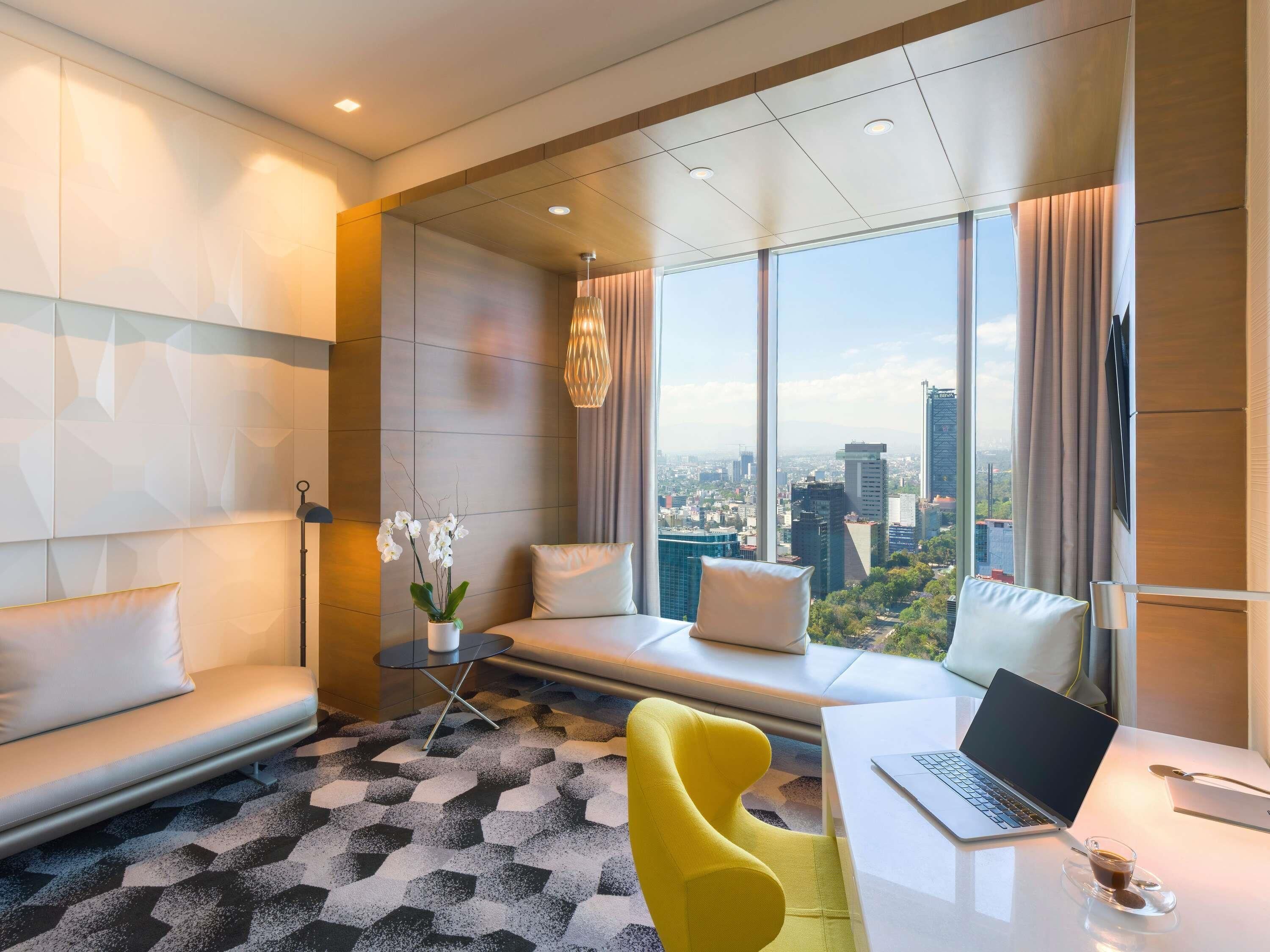 A modern hotel room with a large window, a sofa, a yellow chair, a table with a laptop, and a small table with a cup.