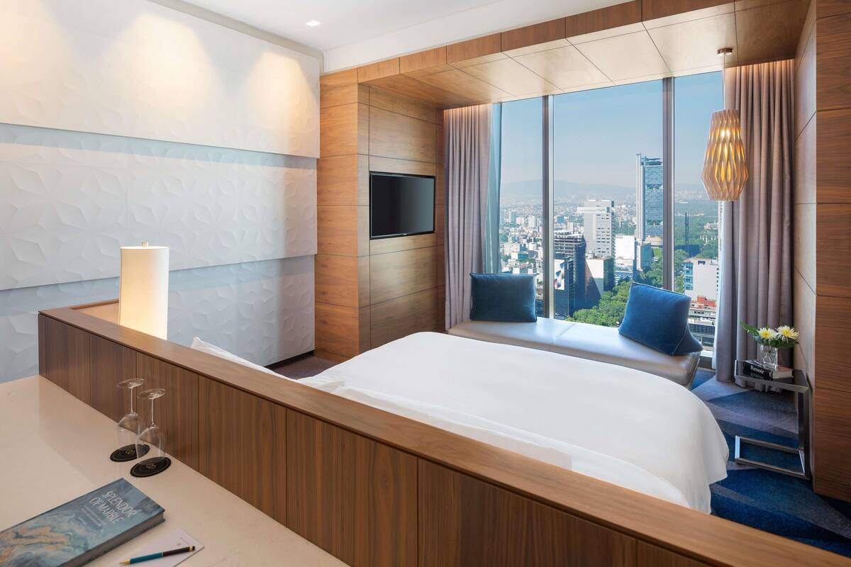 Une chambre d’hôtel moderne dotée d’un grand lit, d’un bureau avec une lampe, d’une télévision et offrant une vue sur la ville.