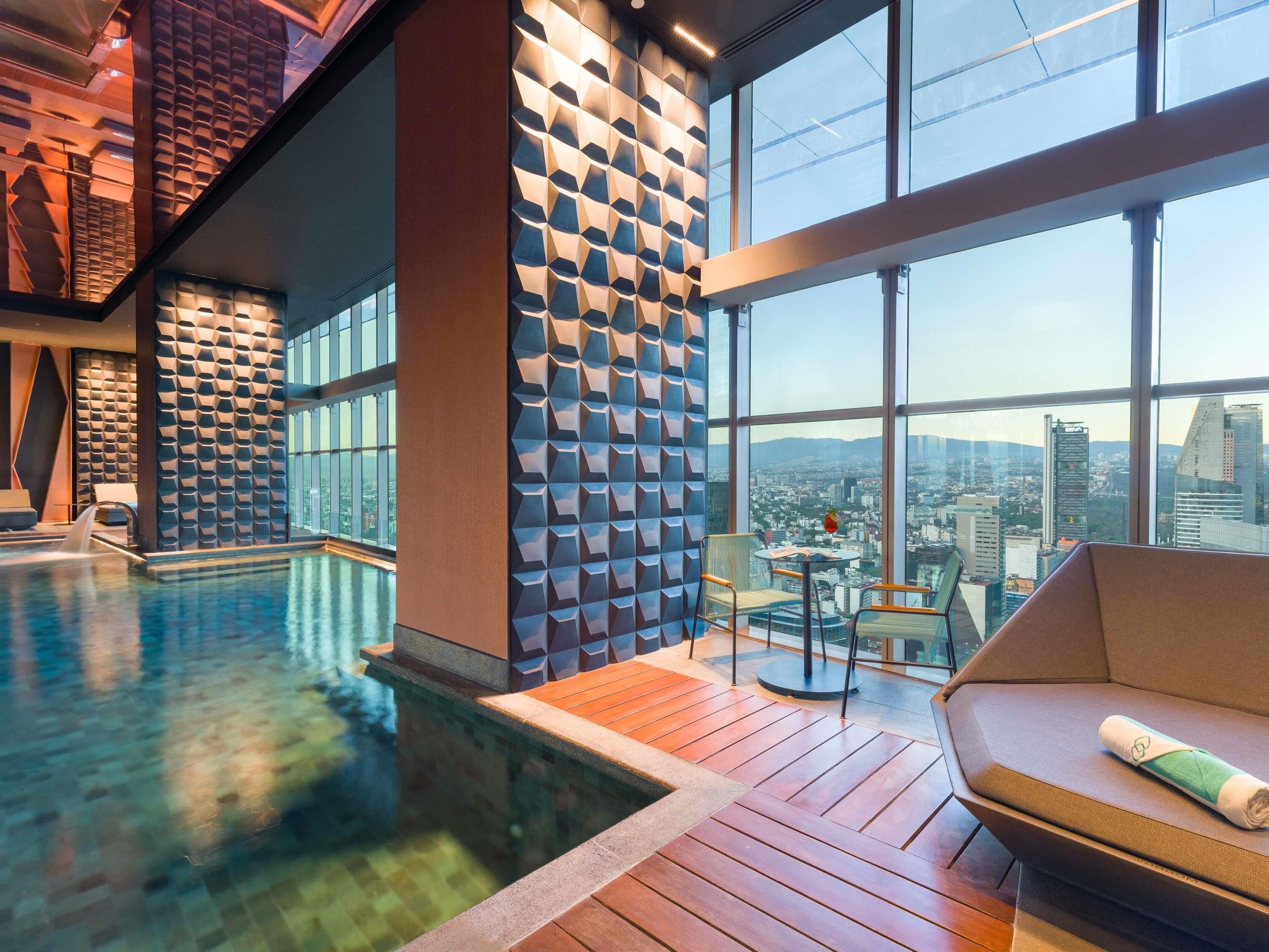 A modern indoor pool area with a city view, a lounge with a sofa, and a unique geometric wall design.