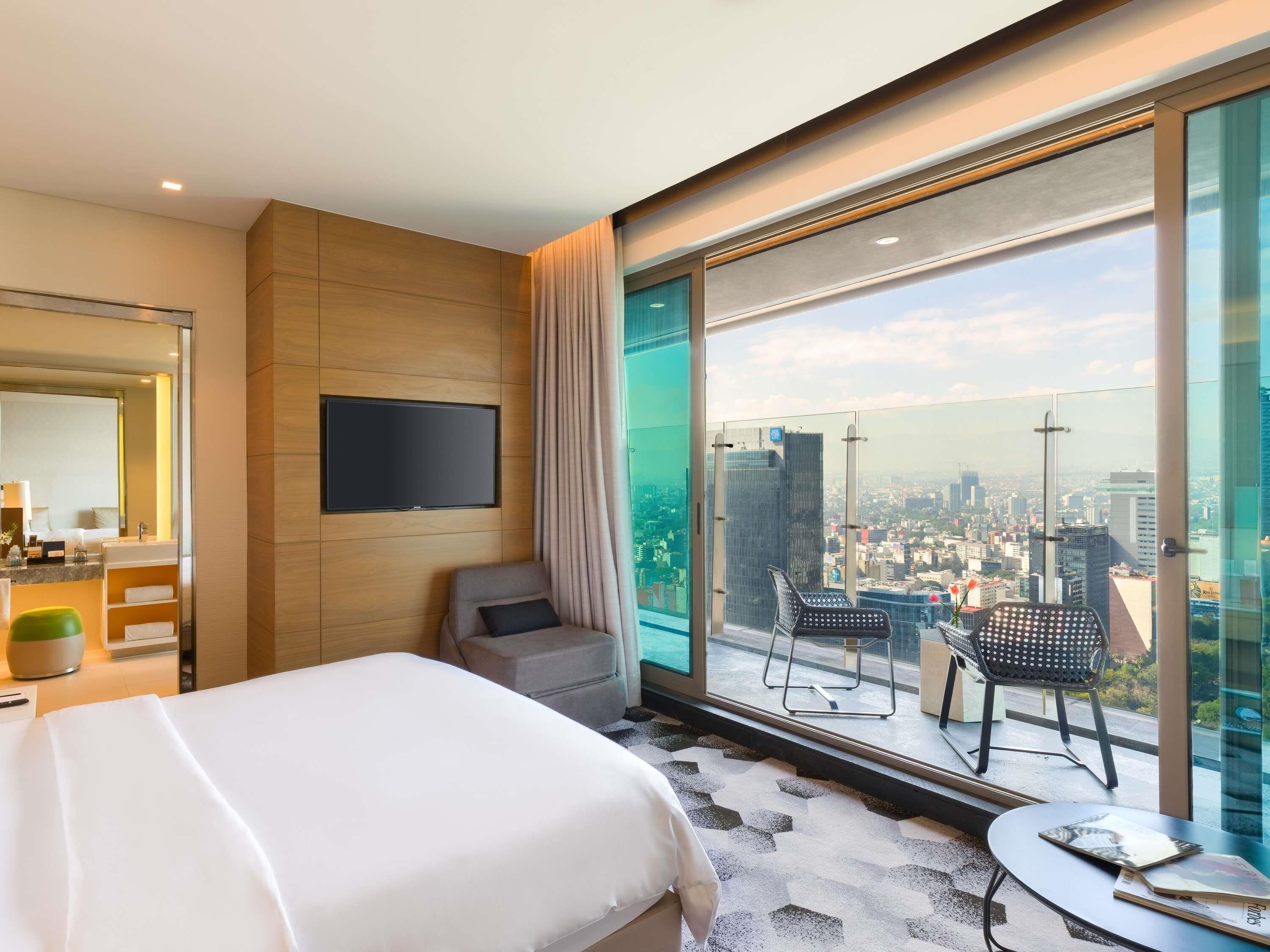Ein Hotelzimmer mit Bett, Fernseher, Badezimmer, großem Fenster mit Blick auf die Stadt und einem Balkon mit Stühlen.
