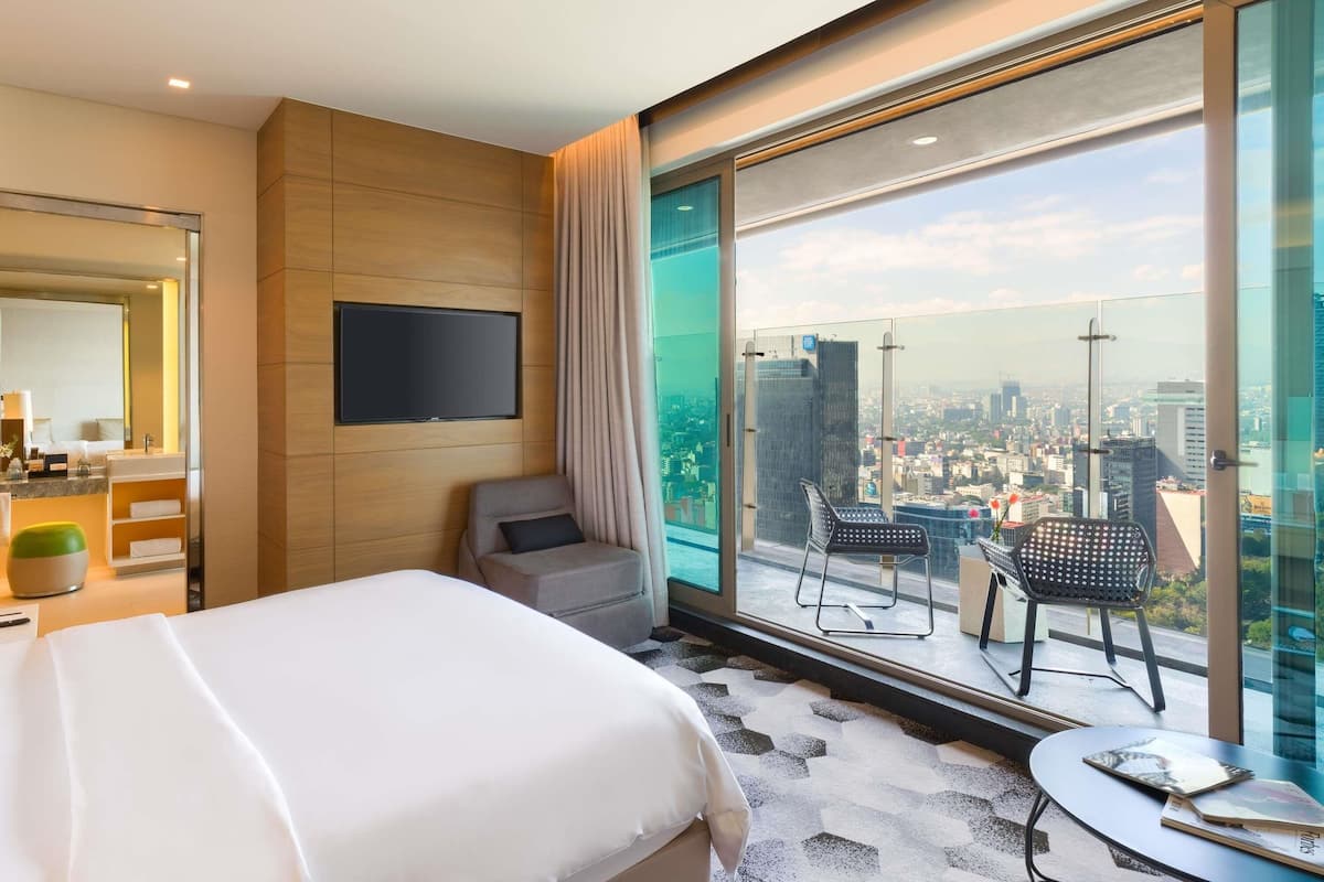 Une chambre d’hôtel comprenant un lit, une télévision, une salle de bain, une grande fenêtre donnant sur la ville et un balcon avec des chaises.