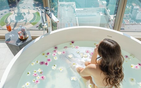 A woman relaxing in a bathtub filled with water and flowers, with a cityscape view through large windows.