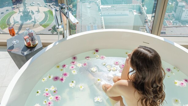 Una mujer se relaja en una bañera llena de agua y flores, con vistas a un paisaje urbano a través de amplias ventanas.