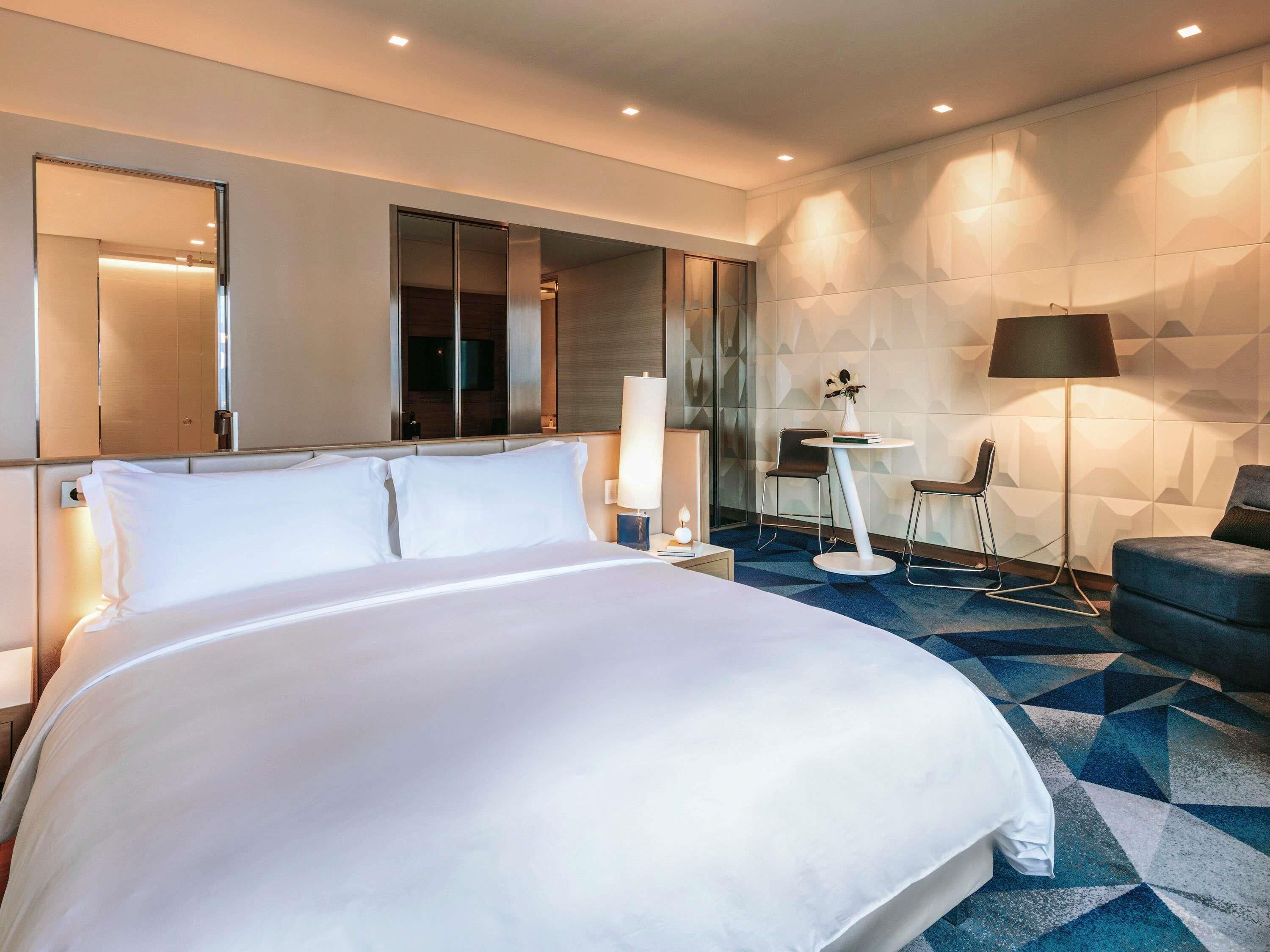 Quarto de hotel moderno, com uma cama grande, uma escrivaninha com cadeira, abajur e espelho.