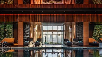 A modern indoor pool area with a clear view of the cityscape through large windows, wooden paneling, and lounge seating.
