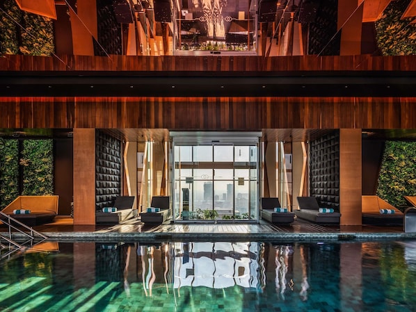 A modern indoor pool area with a clear view of the cityscape through large windows, wooden paneling, and lounge seating.
