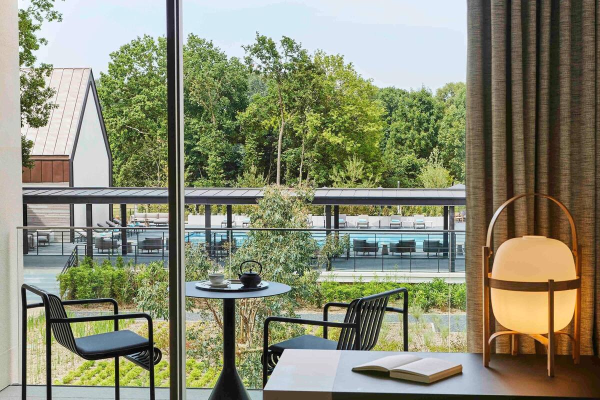 Ein modernes Zimmer mit Blick auf einen Pool und Grünflächen. Es beinhaltet einen Tisch mit Stühlen, eine Lampe und ein Buch.