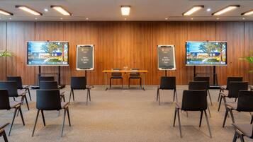 Un salón de conferencias con pared de paneles de madera, dos pantallas grandes, una mesa con sillas y suelo alfombrado.