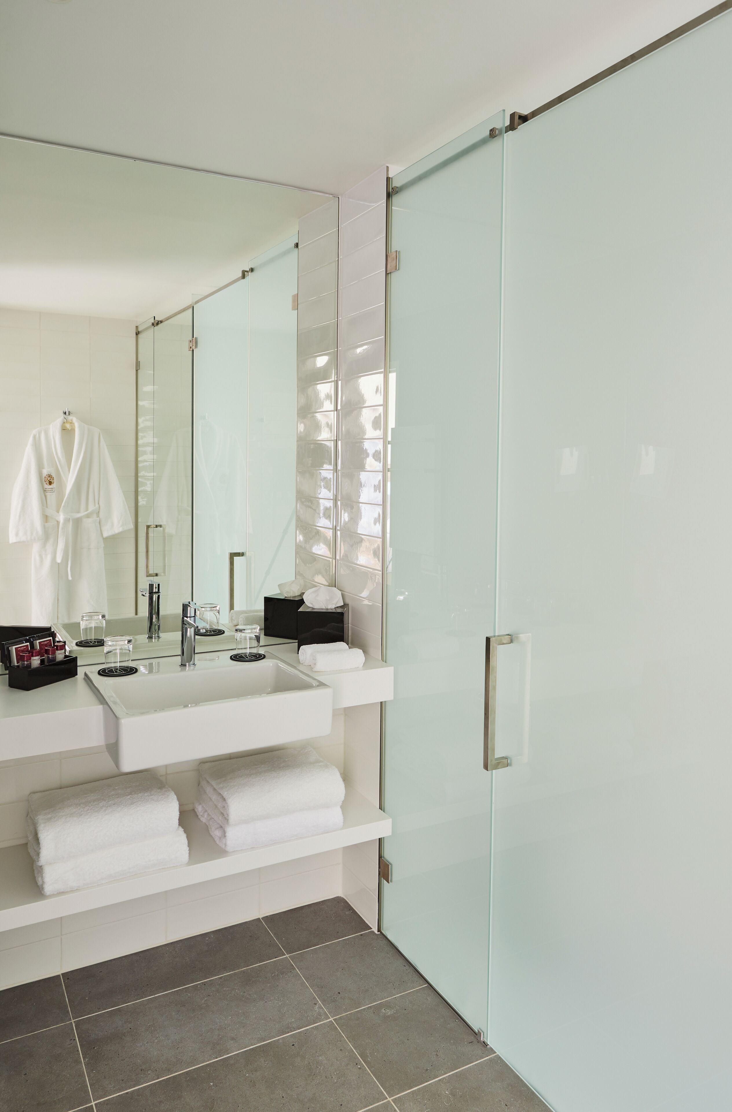 Un baño moderno con un espejo grande, un lavabo blanco, una cabina de ducha de vidrio y toallas dobladas con cuidado en un estante.