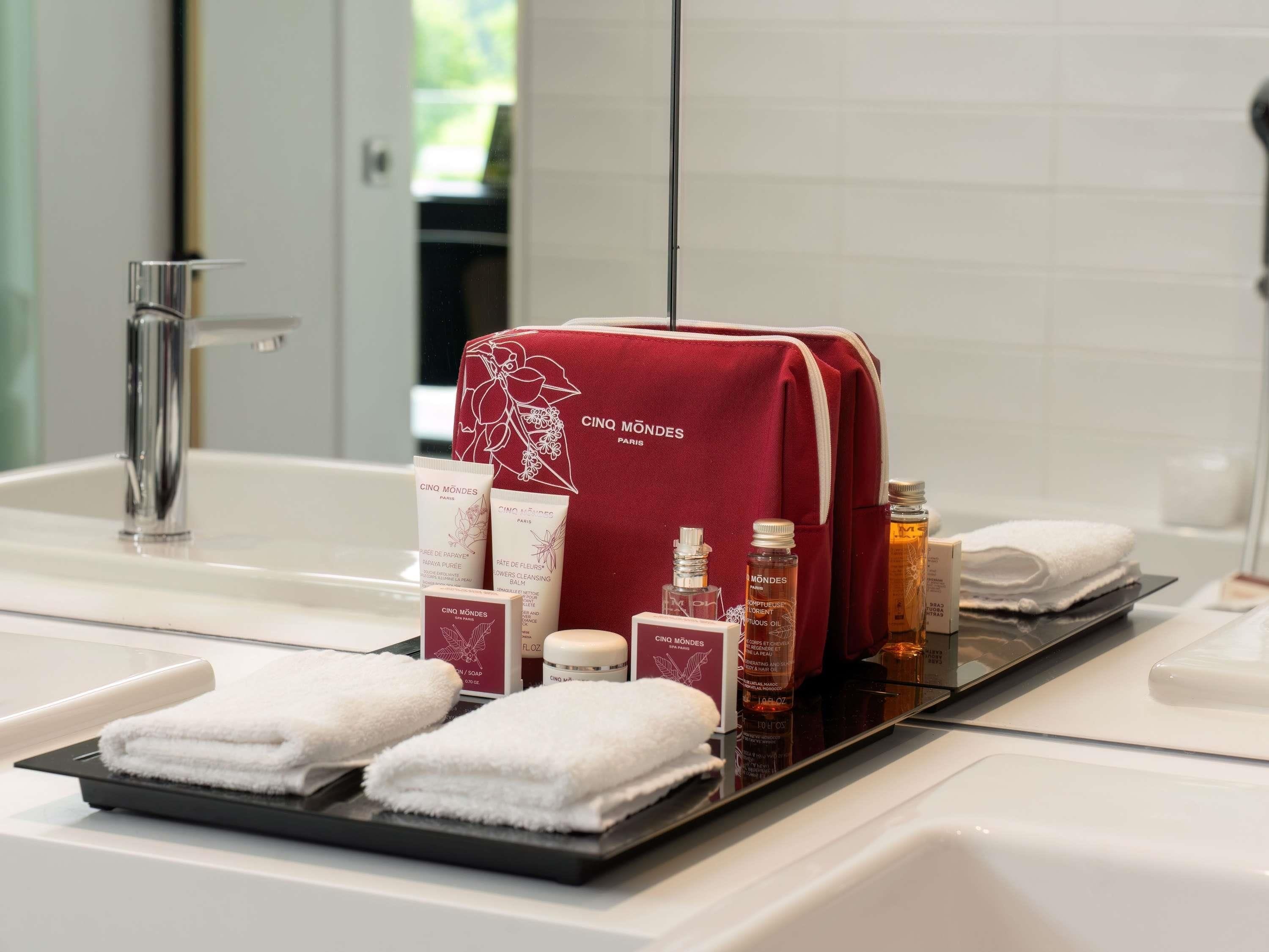 Un baño con lavamanos, espejo y artículos de aseo, incluyendo una bolsa roja, botellas y toallas.