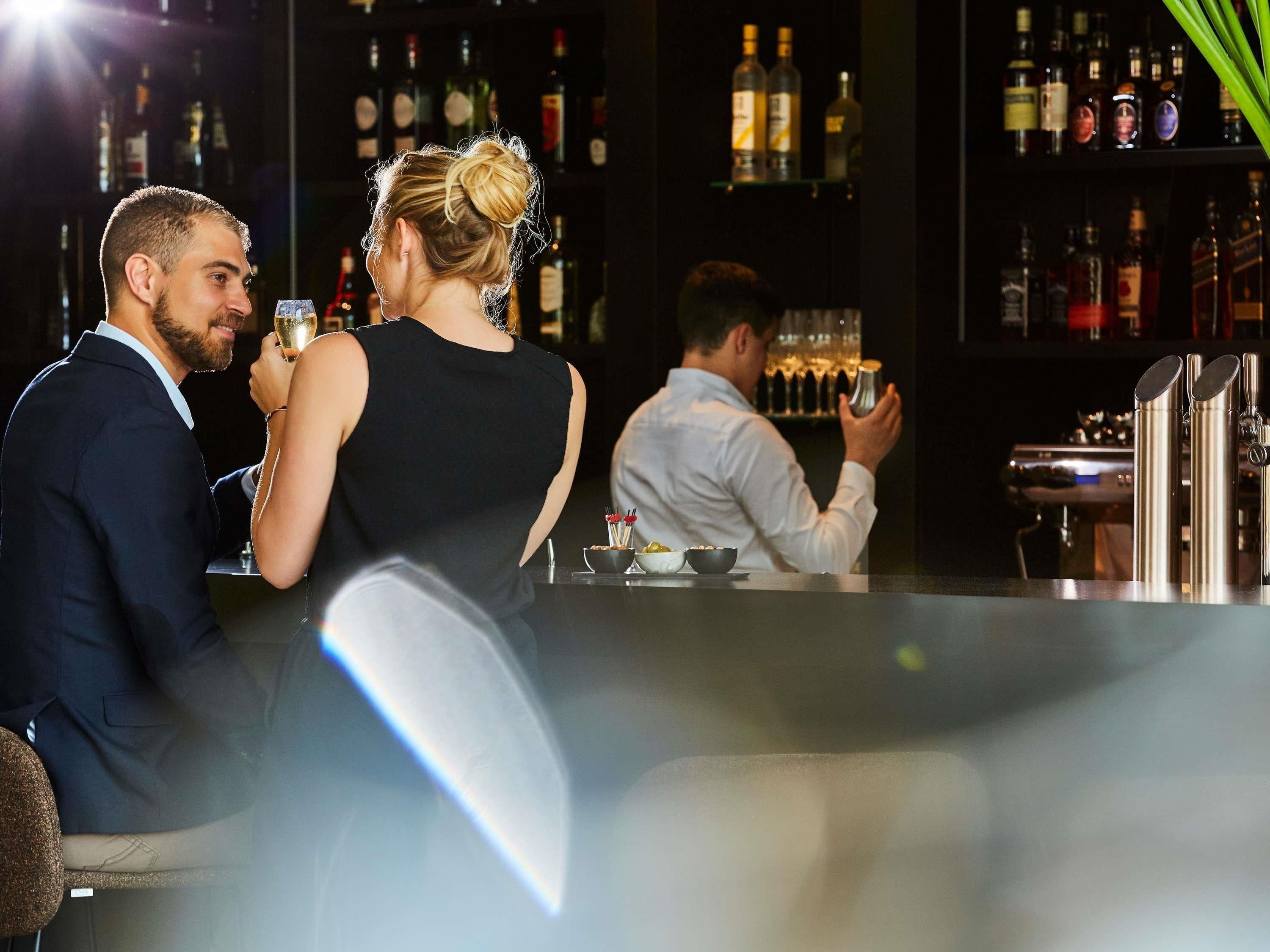 Un bar con un cantinero preparando bebidas, una pareja sentada en una mesa y una variedad de botellas al fondo.
