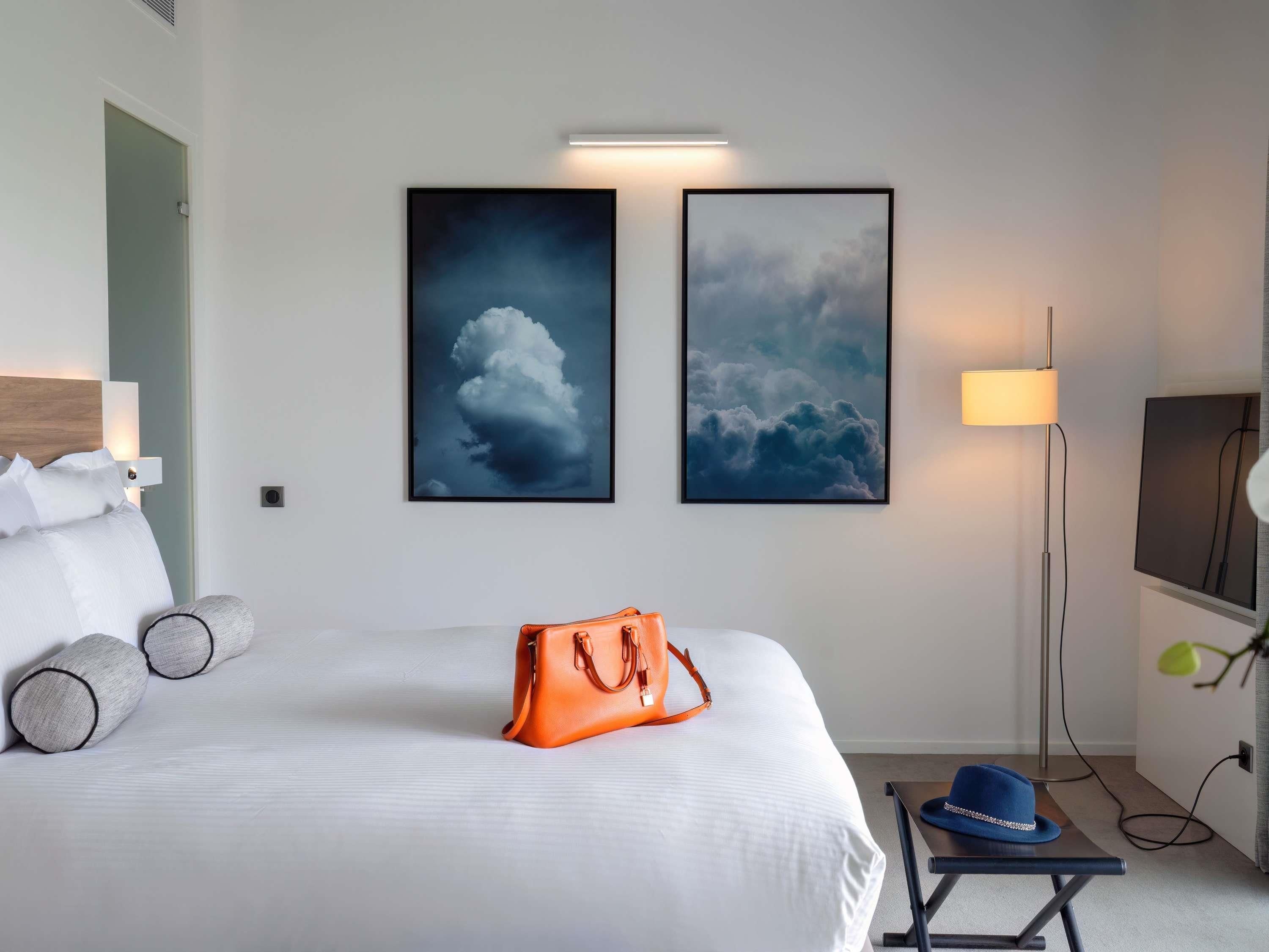 A hotel room with a bed, two framed cloud paintings, a bedside lamp, a table with a hat, and a handbag on the bed.