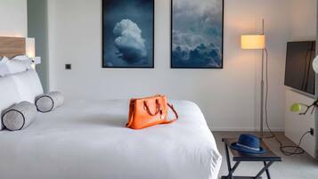 A hotel room with a bed, two framed cloud paintings, a bedside lamp, a table with a hat, and a handbag on the bed.