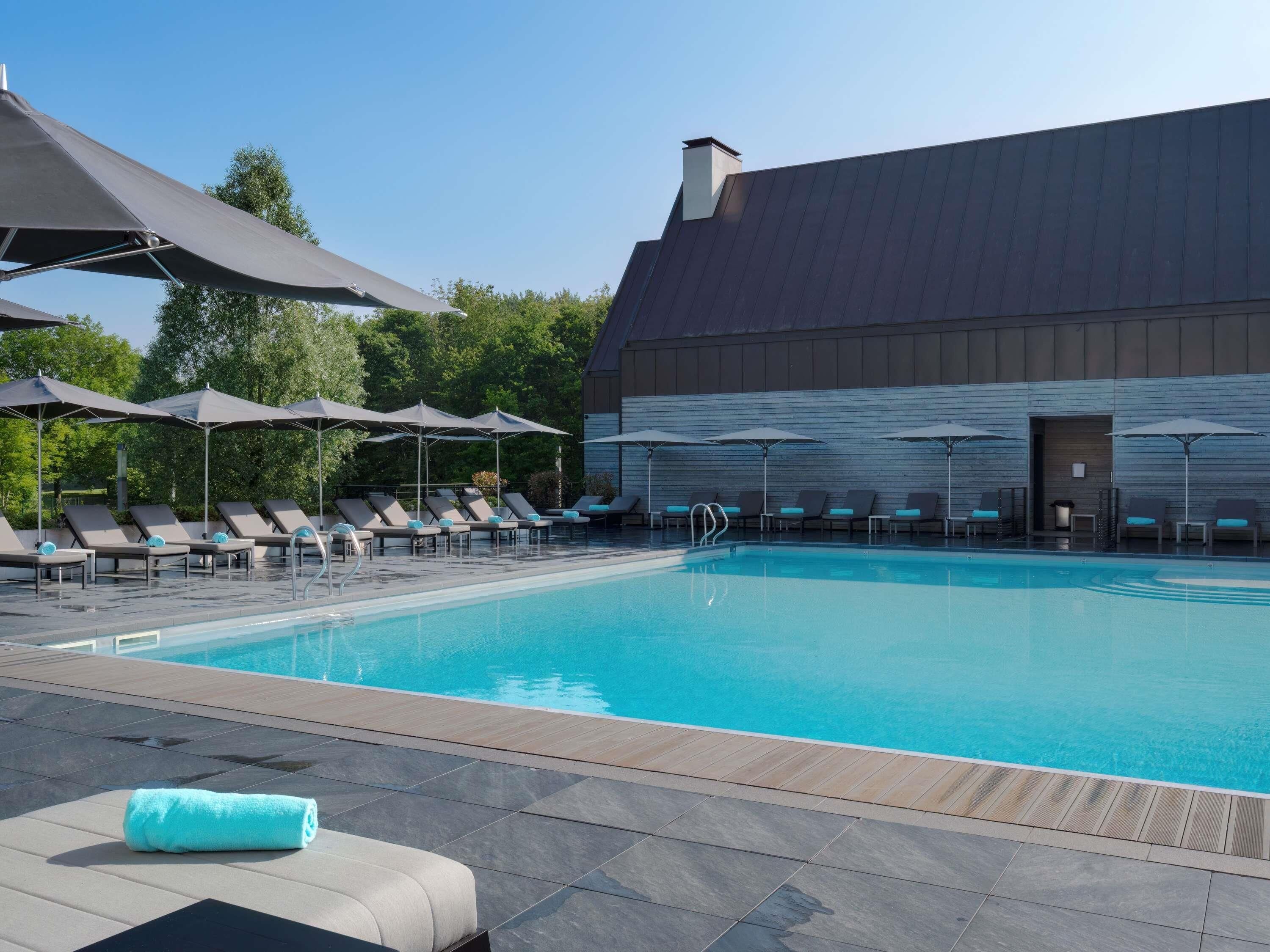 A swimming pool with lounge chairs and umbrellas, a wooden deck, and a building with a dark roof.