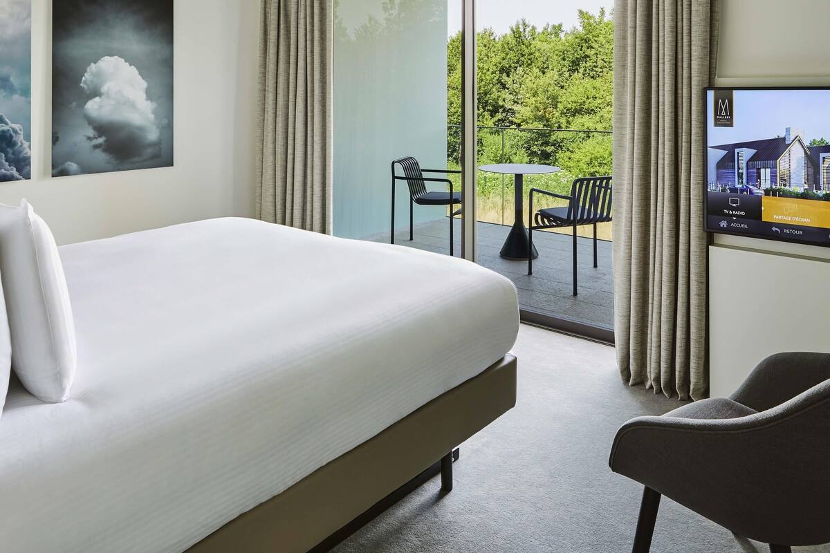 Ein Hotelzimmer mit Bett, Sessel, Tisch und Fernseher, der ein Gebäude zeigt.