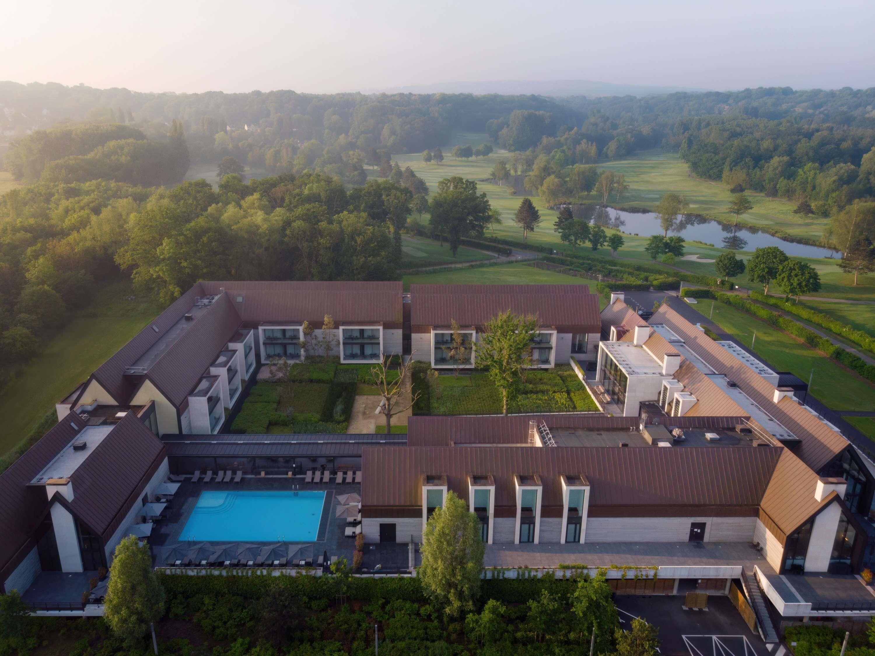A modern hotel with a swimming pool, multiple buildings, and a golf course in the background.