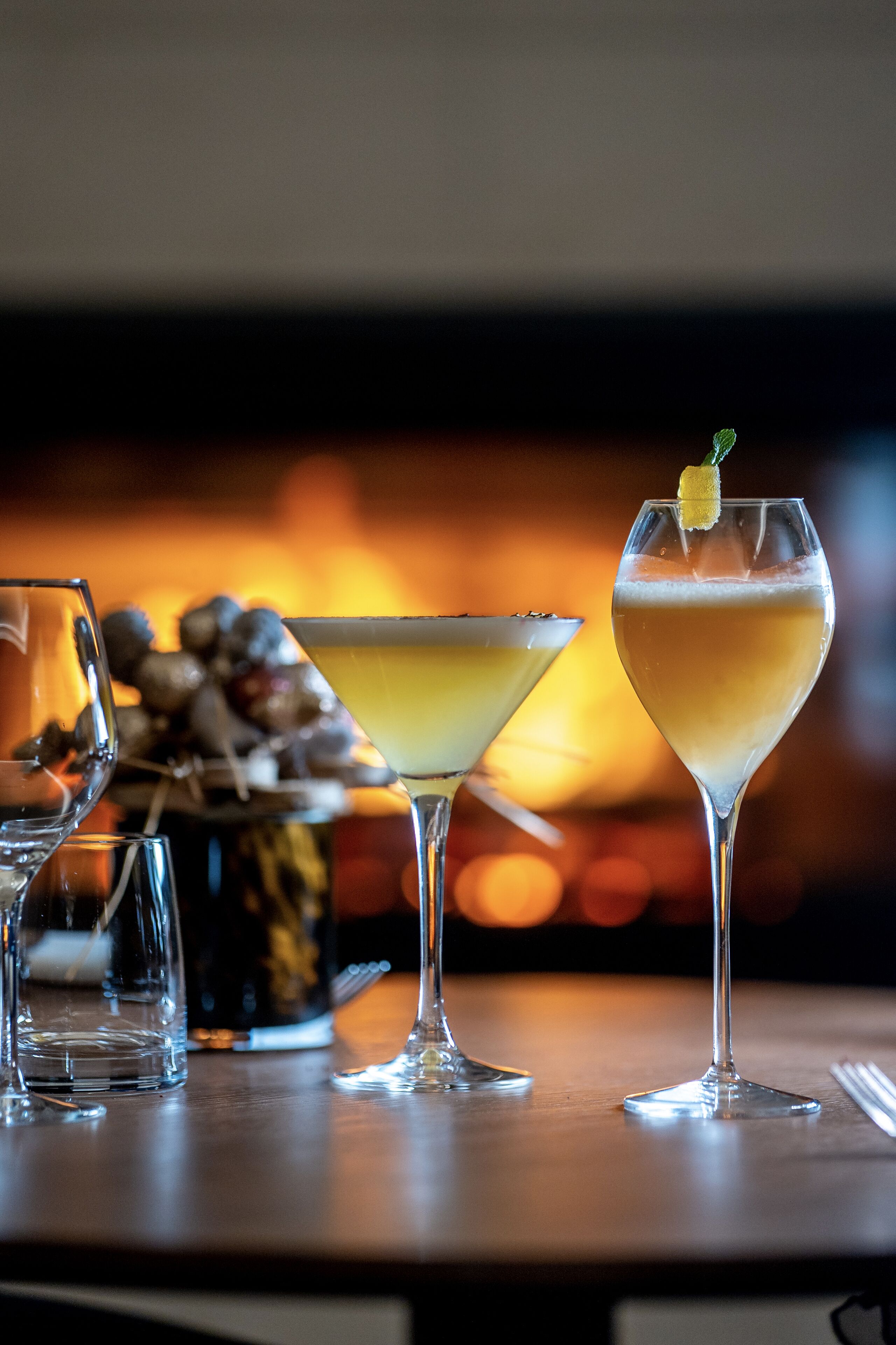 Tres vasos de bebidas sobre una mesa, con un fondo desenfocado de una chimenea.