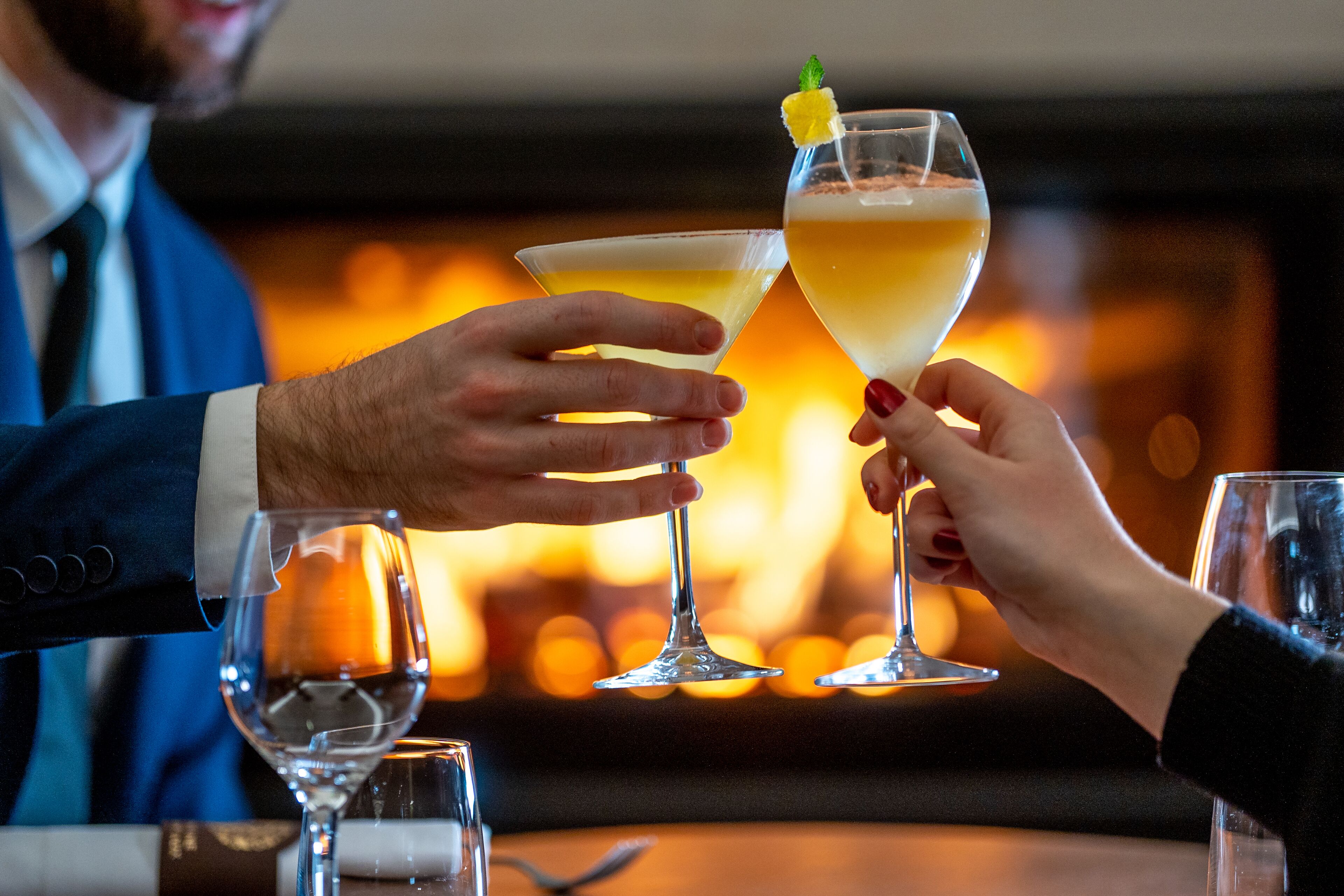 Two people toasting with champagne glasses in a warmly lit room with a fireplace in the background.