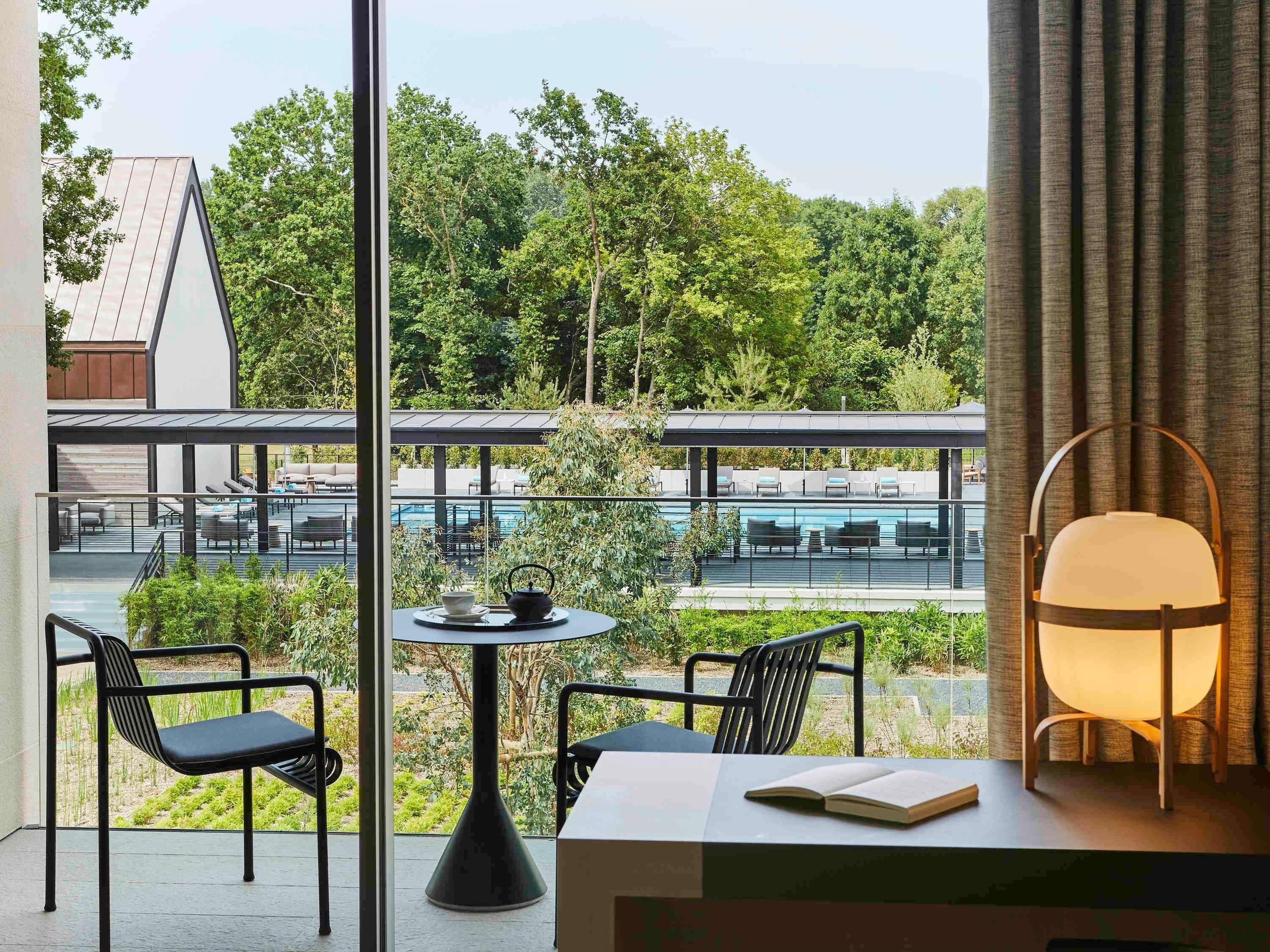 A modern room with a view of a pool and greenery. It includes a table with chairs, a lamp, and a book.