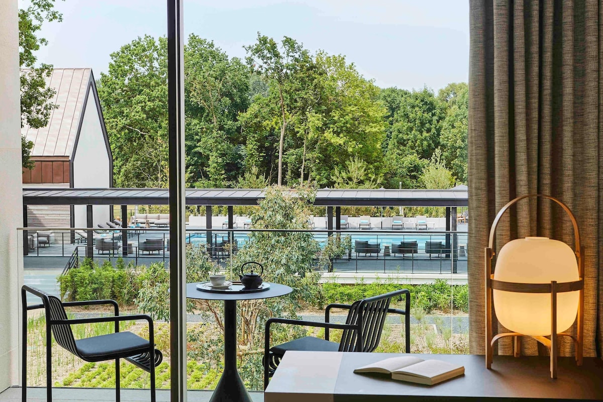 A modern room with a view of a pool and greenery. It includes a table with chairs, a lamp, and a book.