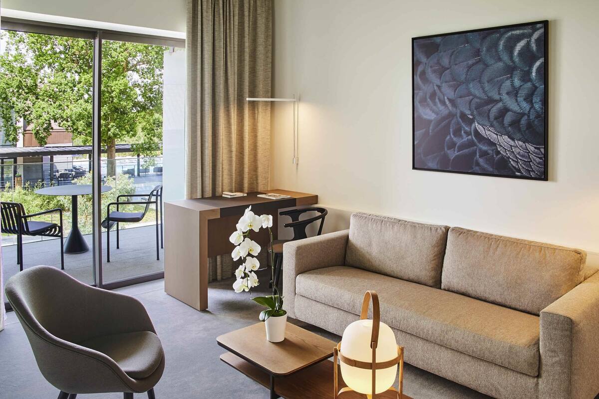 Ein modernes Wohnzimmer mit einer Couch, einem Sessel, einem Couchtisch mit Lampe, einem Schreibtisch und einem großen Fenster mit Vorhängen.