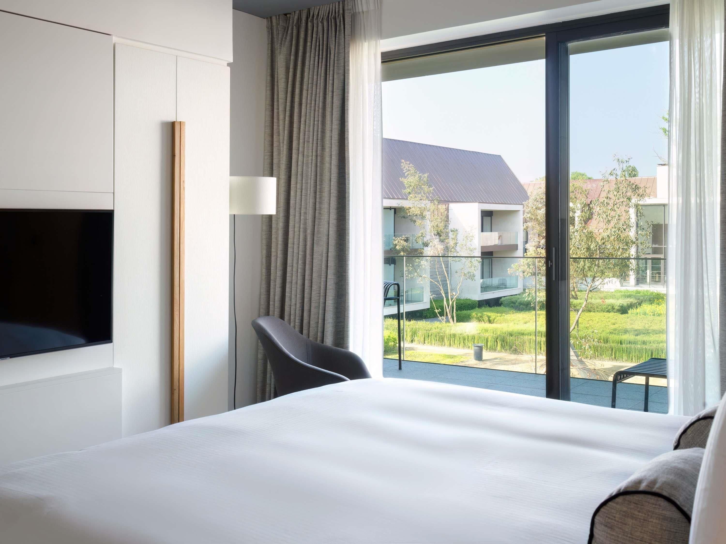 Une chambre d’hôtel avec un lit, une télévision, une chaise et un balcon donnant sur un immeuble et des espaces verts.