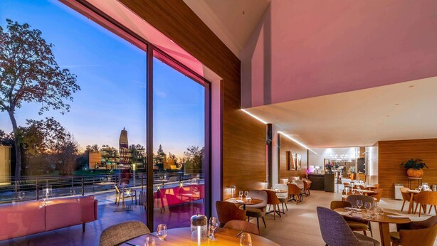 A restaurant with a view of a cityscape, featuring a large dining area with round tables and chairs, a bar area, and a seating area with a view of the outdoors. - Le Domaine des Vanneaux Golf & Spa L'Isle Adam - MGallery Collection (Presles)