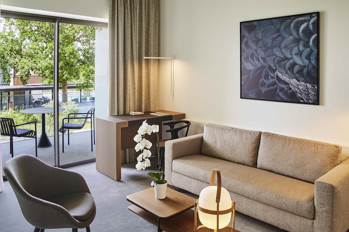 A modern living room with a sofa, a chair, a coffee table with a lamp, a desk, and a large window with curtains.