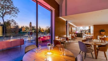 A restaurant with a view of a cityscape, featuring a large dining area with round tables and chairs, a bar area, and a seating area with a view of the outdoors.