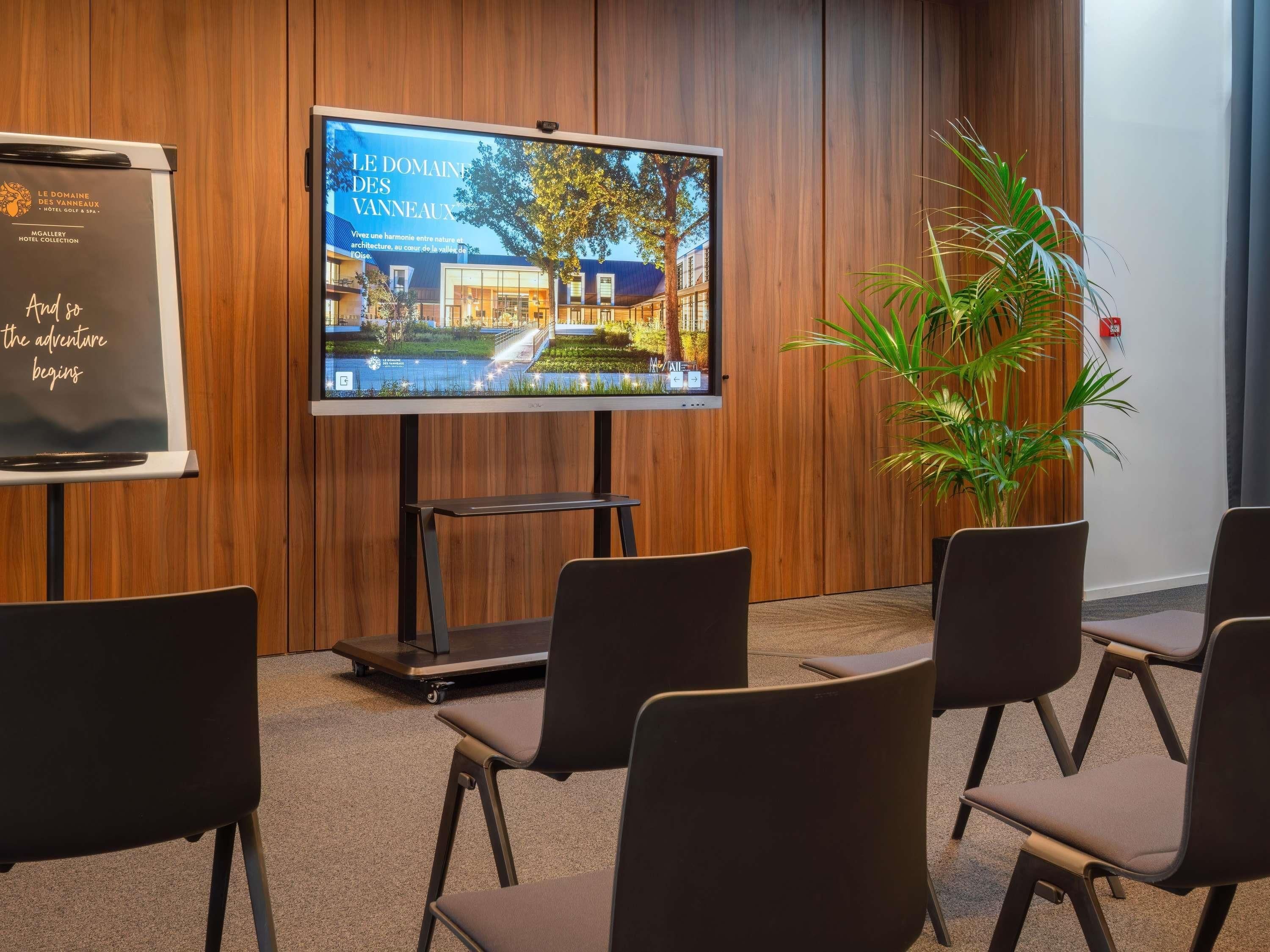 A conference room with a large screen displaying a hotel interior, wooden panel walls, and black chairs.