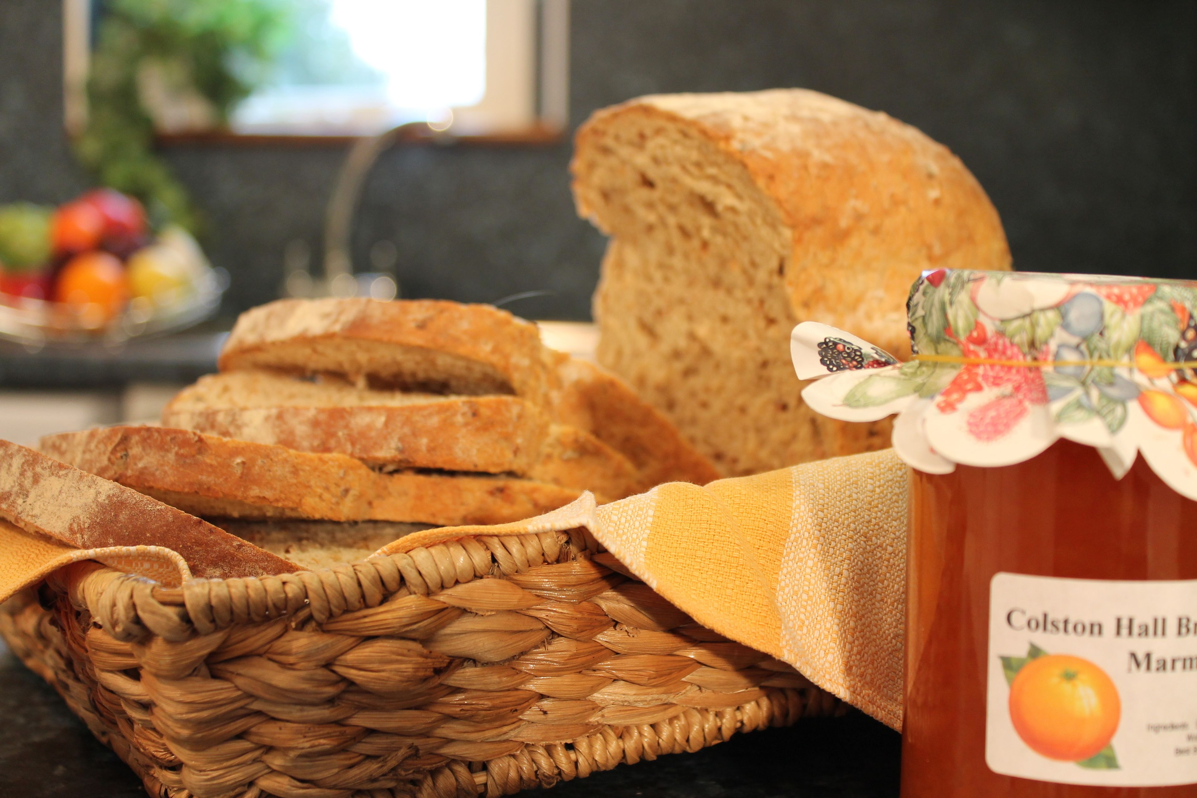 A basket of bread, a jar of marmalade, and a plate of fruit in a kitchen setting.