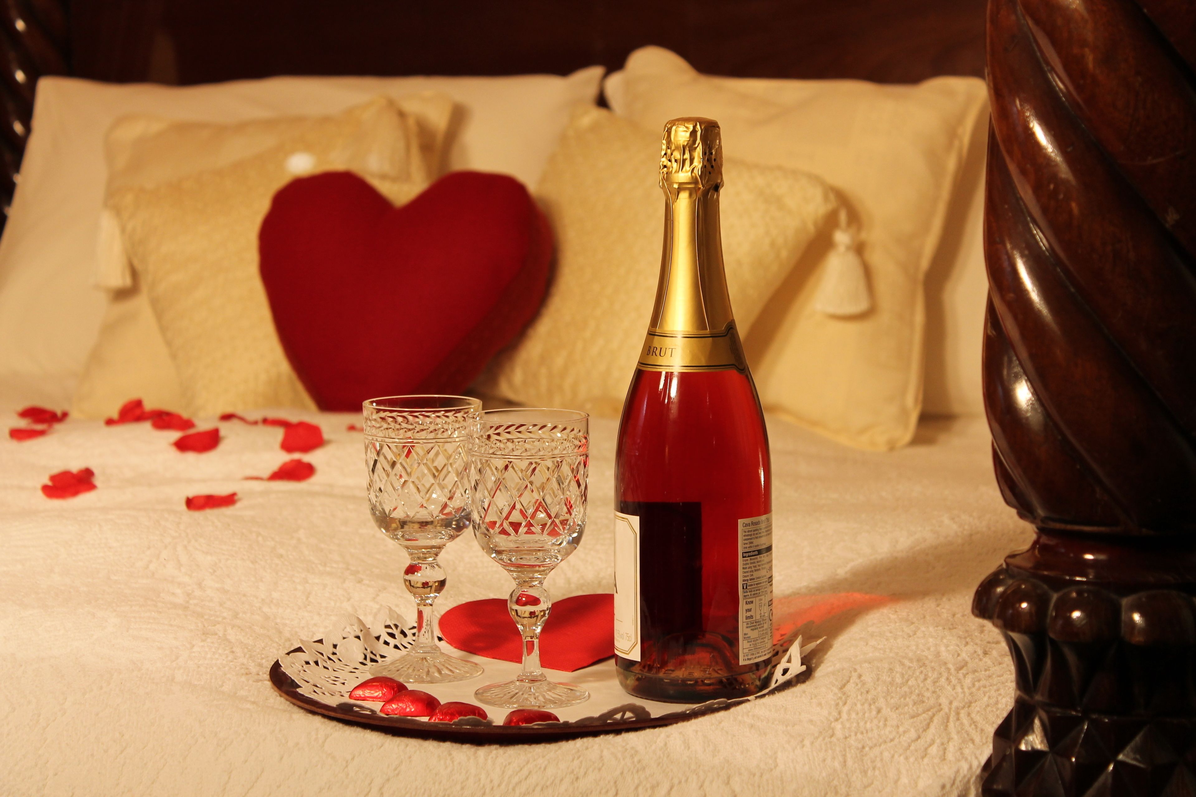 A bed with a bottle of champagne, two glasses, and rose petals on a doily.