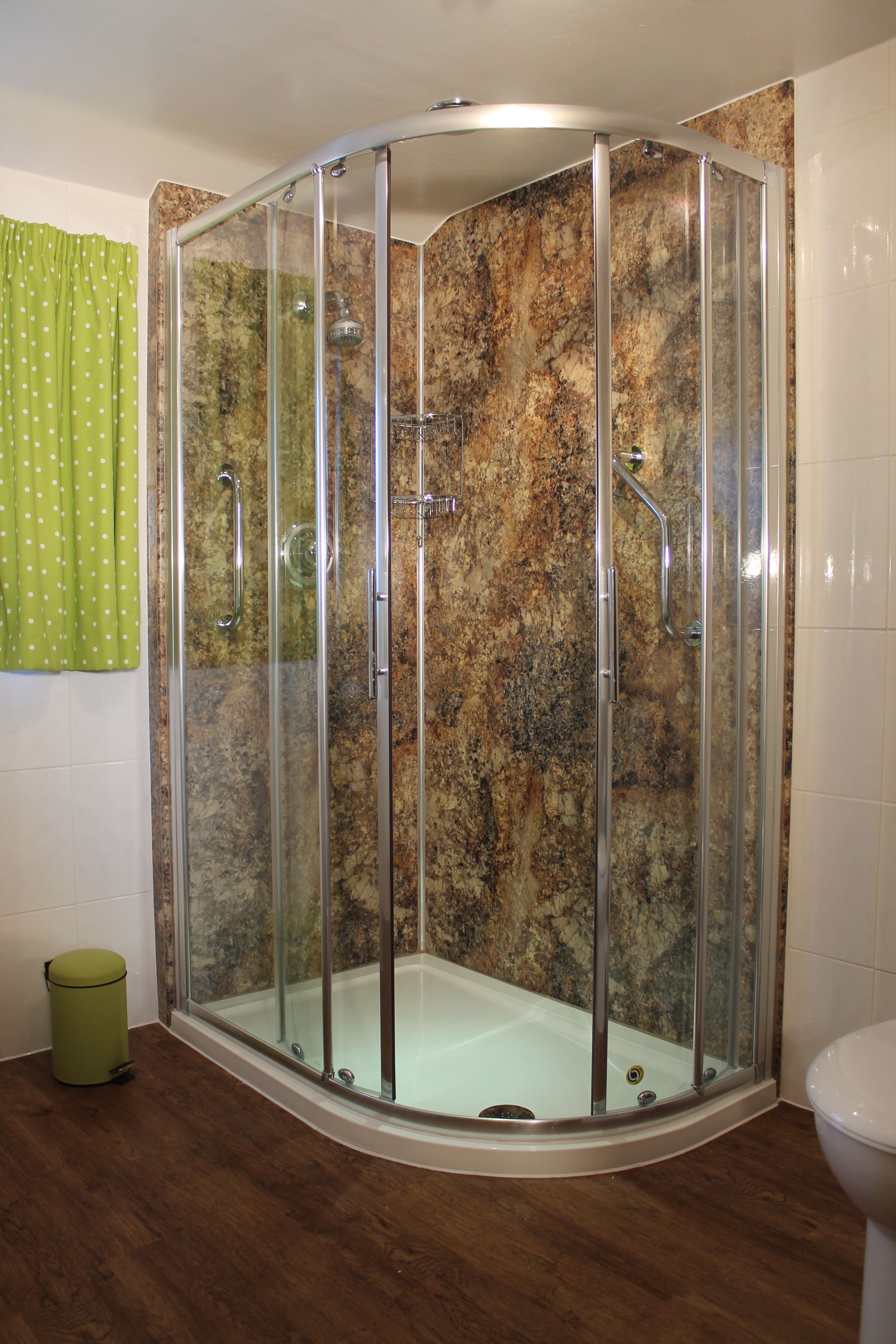 A glass-enclosed shower with a curved design, a bathtub, and a toilet in a bathroom with wooden flooring and a green curtain.