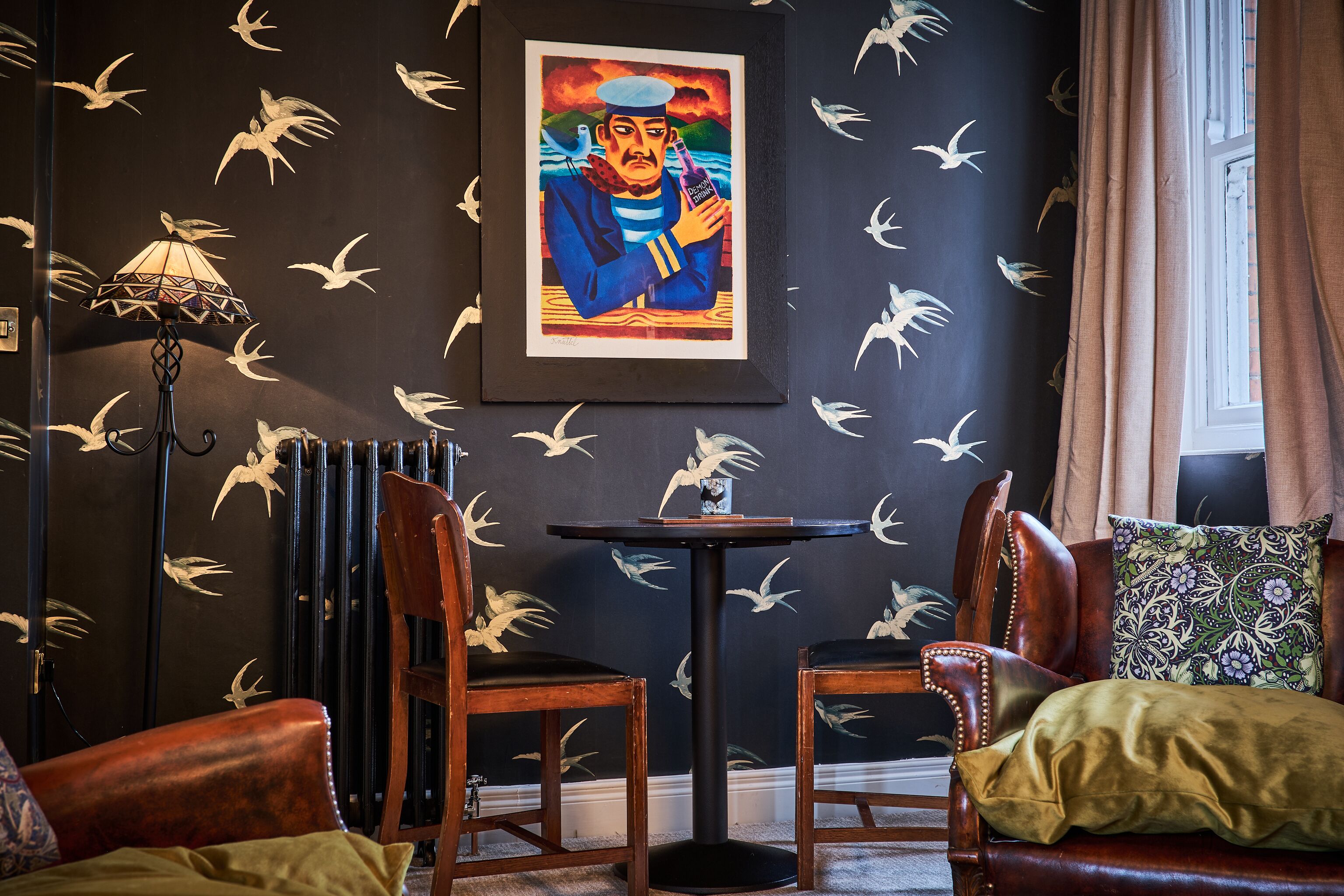 Una habitación con una pared oscura con diseño de pájaros, una obra de arte enmarcada, una mesa, dos sillas, una lámpara y una ventana con cortinas.