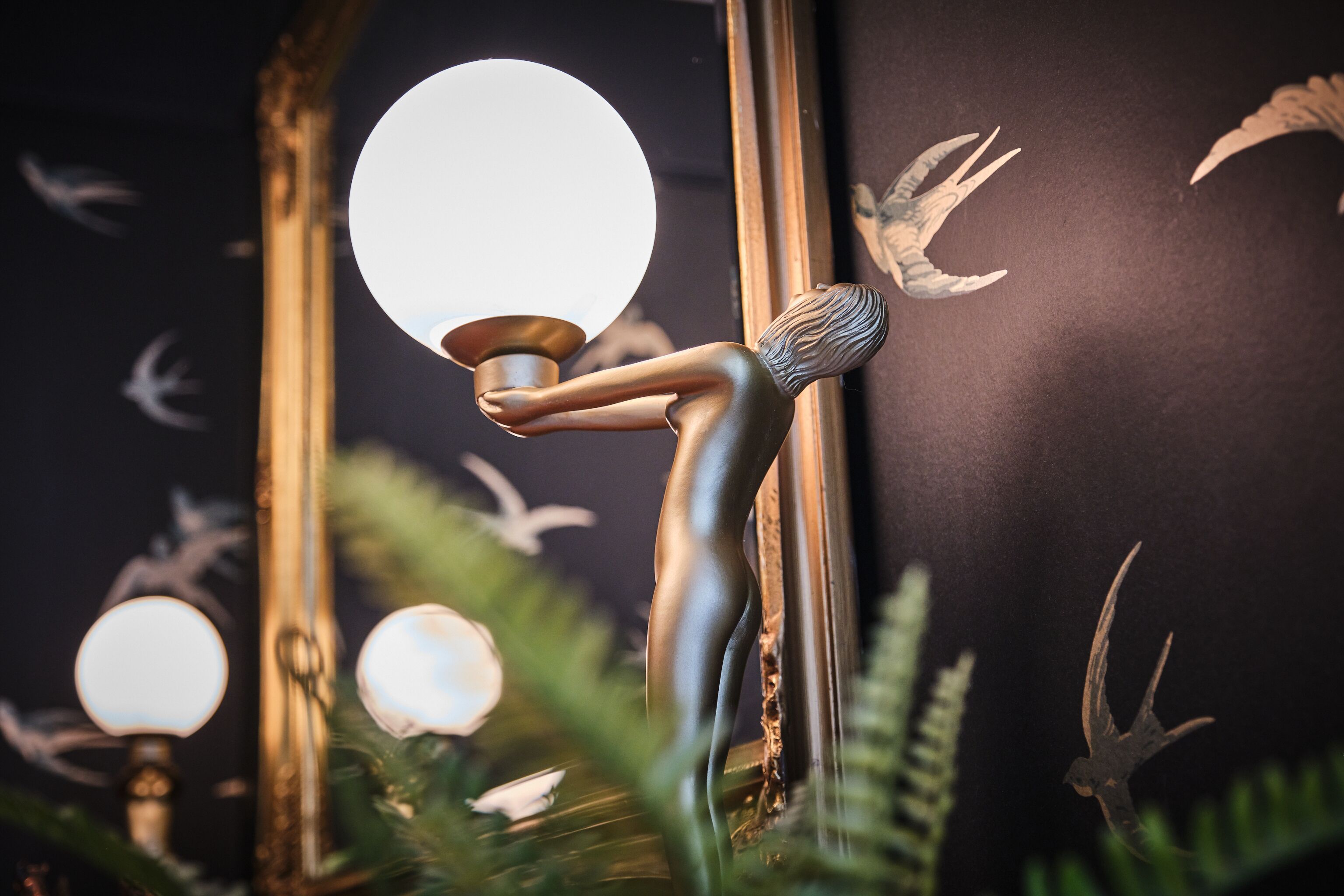 En skulptur af en figur der holder en lysarmatur, med et spejl der reflekterer en fugl og en rund lyskilde.