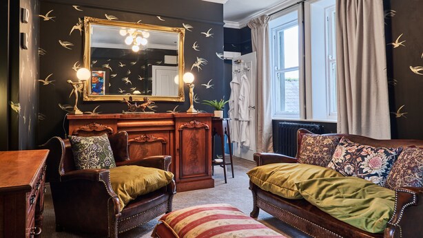 Una habitación con un espejo grande, una tocador con luces, un sofá con cojines estampados y vistas al exterior.