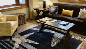 Ein Wohnzimmer mit einer Couch, Sesseln, einem Couchtisch und einer Lampe. Es gibt Fenster mit Vorhängen, einen Teppich und einen Deckenventilator.