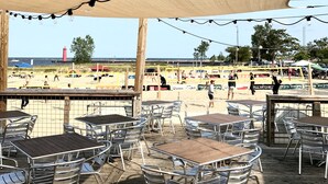 Ein Sitzbereich im Freien mit Tischen und Stühlen, ein Beachvolleyballnetz und Blick auf den Strand und den Leuchtturm.