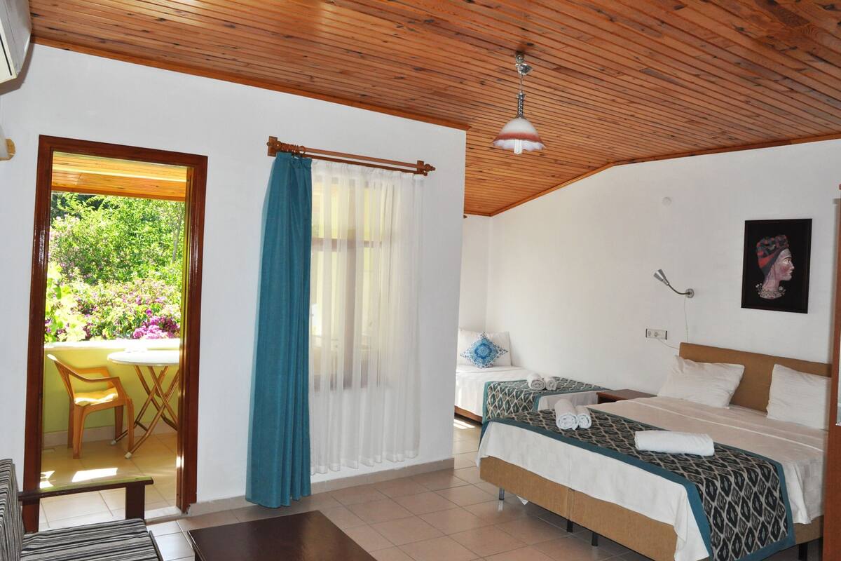 A bedroom with a bed, a bedside table, a hanging lamp, a window with curtains, and a view of greenery outside.