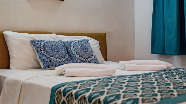 A neatly made bed with patterned bedding, a framed artwork above it, and a teal curtain on the window. - San Simon Hotel (Kemer)