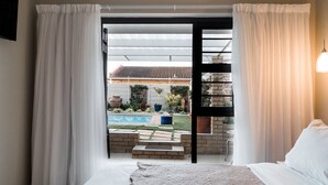 A bedroom with a large bed, white curtains, and a view of a backyard with a pool and outdoor furniture. - Wagtails Guest House (Gqeberha (Port Elizabeth))