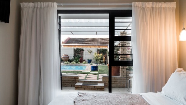 A bedroom with a large bed, white curtains, and a view of a backyard with a pool and outdoor furniture. - Wagtails Guest House (Gqeberha (Port Elizabeth))