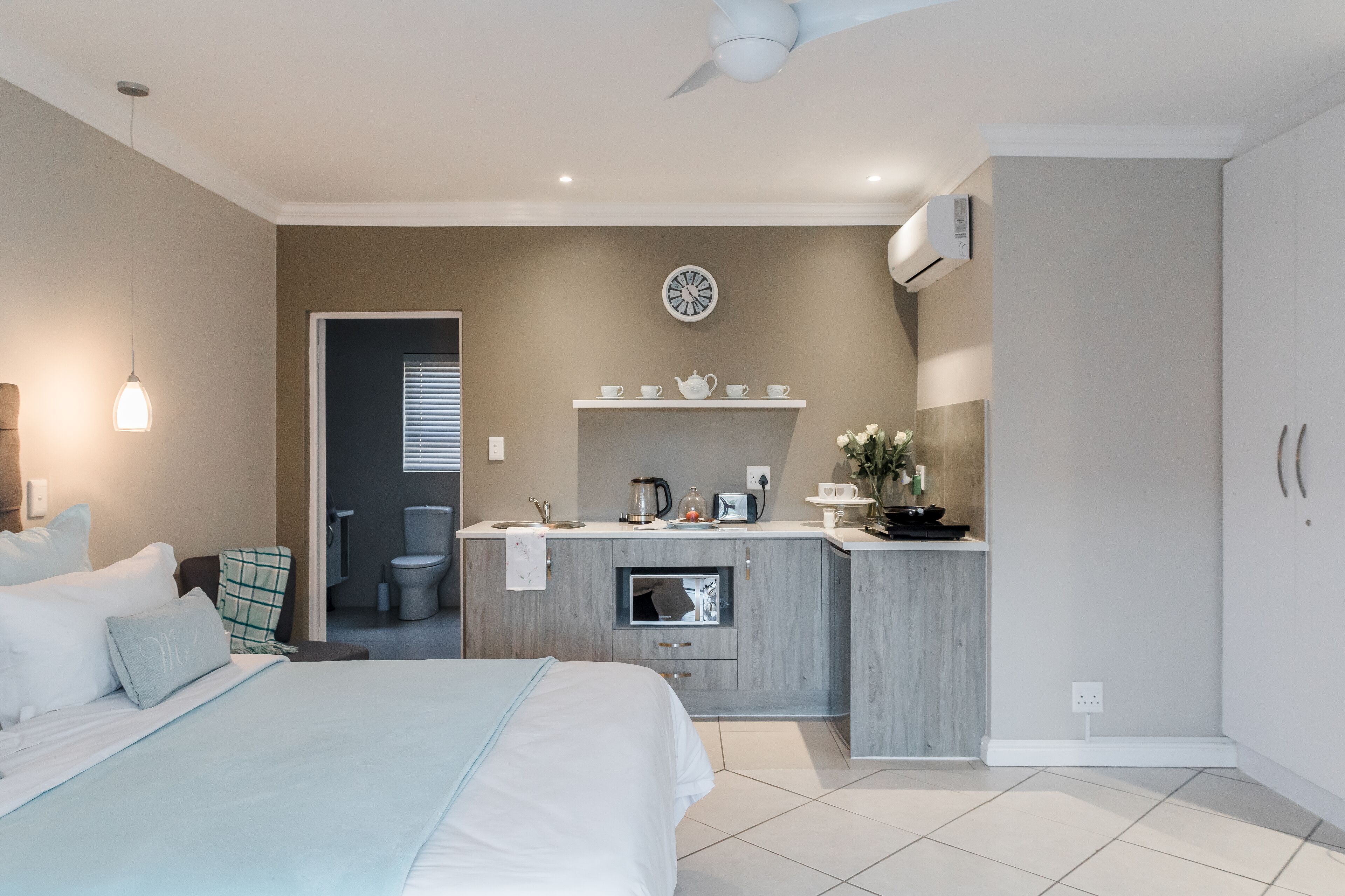 A modern bedroom with a large bed, a built-in kitchenette, a ceiling fan, and a clock on the wall.