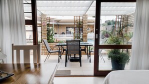 A room with a bed, a wooden table, and a dining area with chairs and a table. There is a view of a brick building and a white fence in the background. - Wagtails Guest House (Gqeberha (Port Elizabeth))
