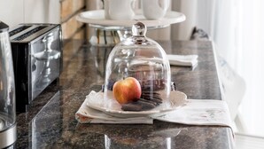 A breakfast setup with a glass dome over an apple and cookies, a white cup, and a vase with flowers on a marble countertop. - Wagtails Guest House (Gqeberha (Port Elizabeth))