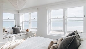Ein Schlafzimmer mit einem großen Bett, einer Sitzgelegenheit am Fenster mit Blick auf den Strand und Meerblick durch mehrere Fenster.