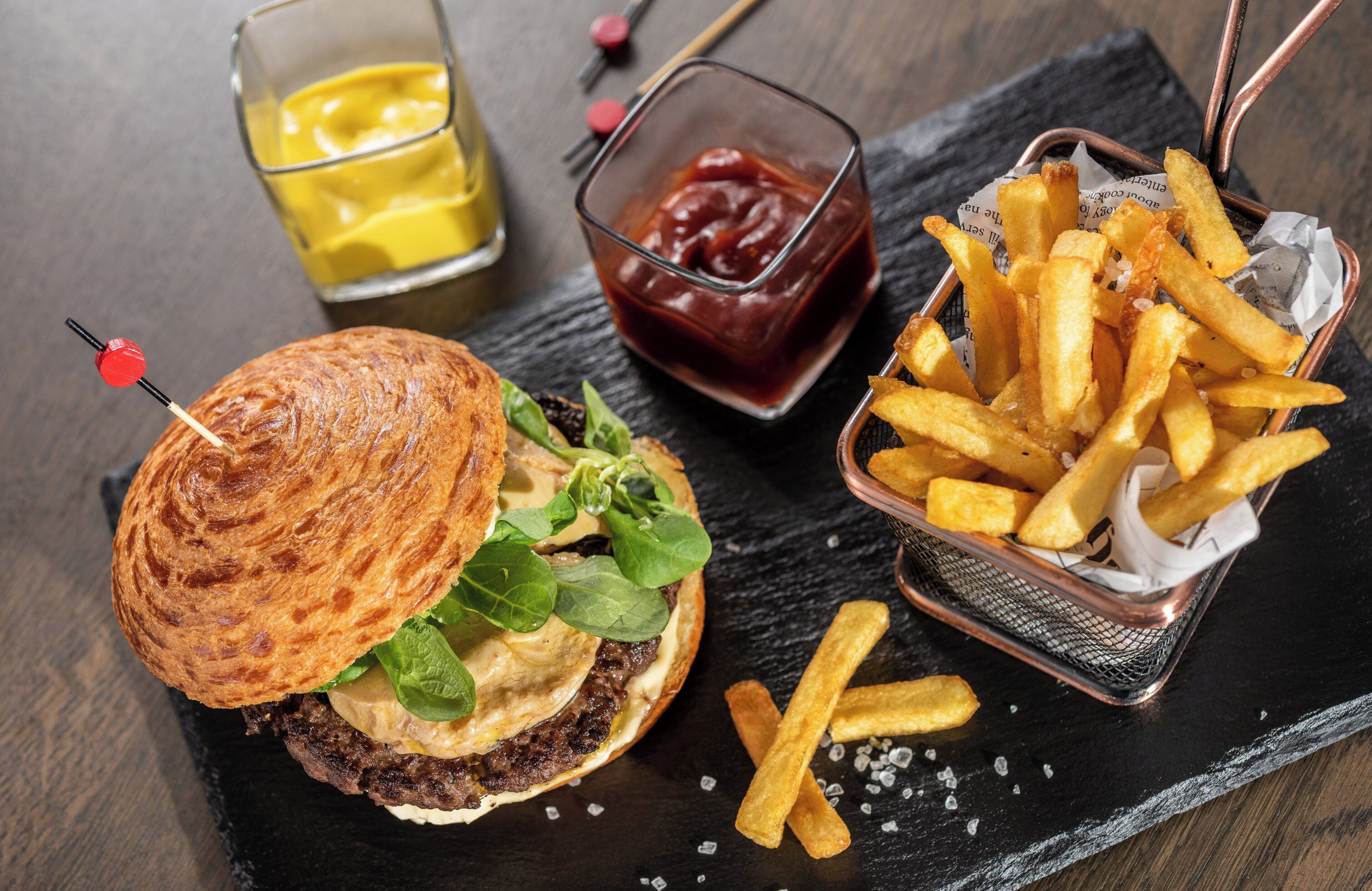 A burger with a toasted bun, lettuce, cheese, and a beef patty, accompanied by French fries in a basket and a glass of mustard.