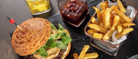 A burger with a toasted bun, lettuce, cheese, and a beef patty, accompanied by French fries in a basket and a glass of mustard.