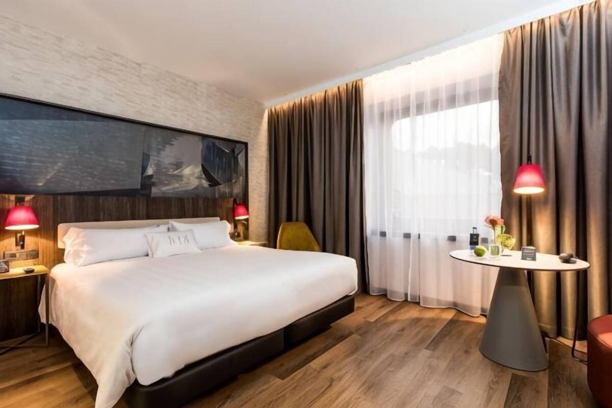 A hotel room with a large bed, a bedside table with lamps, a small round table with a vase, and a view through sheer curtains.